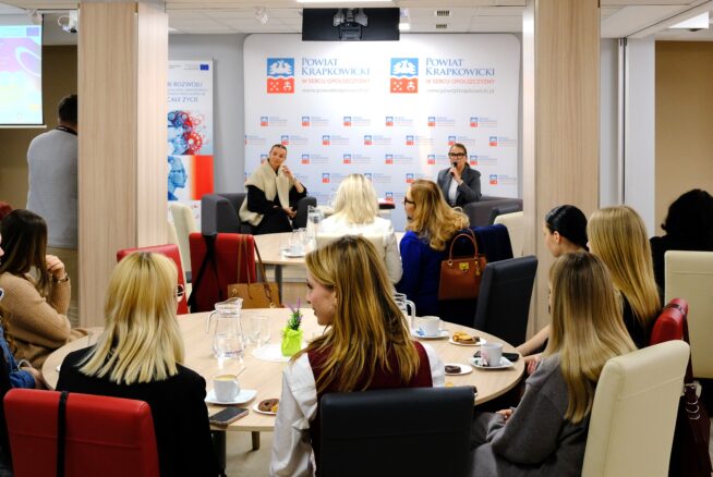 Grupa osób siedzi przy okrągłych stołach w nowoczesnej sali konferencyjnej, słuchając dwóch prelegentów omawiających uczenie się przez całe życie. Banery i flaga Unii Europejskiej są widoczne, gdy uczestnicy robią notatki i angażują się w temat.