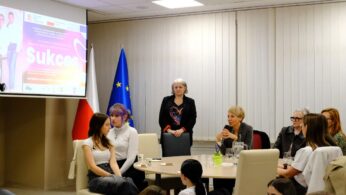 Grupa kobiet siedzi przy stołach i dyskutuje w sali konferencyjnej. Widoczne są dwie flagi (Polski i UE) oraz ekran projektora wyświetlający 