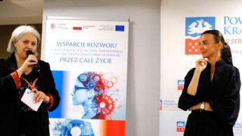 Dwie kobiety przemawiają podczas wydarzenia. Jedna, trzymając mikrofon i dokumenty, zwraca się do publiczności; druga stoi, słuchając uważnie. Za nimi znajduje się baner w języku polskim z grafiką przedstawiającą profil kobiety i koła zębate oraz logo Powiatu Krakowskiego.