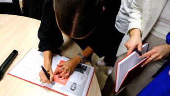 Kobieta z brązowymi włosami związanymi z tyłu podpisuje książkę z czarno-białym zdjęciem i czerwoną okładką przy okrągłym drewnianym stole. Jest otoczona przez inne osoby, z których jedna trzyma długopis, a druga egzemplarz książki. Widoczne są tylko ich ręce i tułowia.