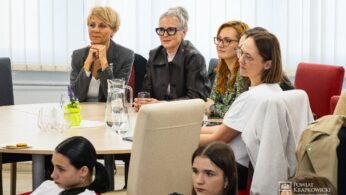 Grupa kobiet siedzi uważnie przy stole w jasnym pokoju z dużymi oknami. Niektóre z nich uśmiechają się lub uważnie słuchają. Na stole stoją dzbanki z wodą, szklanki i mały wazon z kwiatami. Atmosfera wydaje się profesjonalna i wciągająca.