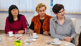 Trzy kobiety siedzą przy stole zastawionym białymi filiżankami, spodkami, szklankami, dzbankiem z wodą i żółtym kwiatem. Są uśmiechnięte i zaangażowane, a światło z pobliskich okien oświetla scenę.