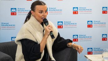 Kobieta z zaczesanymi do tyłu włosami mówi do mikrofonu, siedząc. Ma na sobie kremową, rozmytą kamizelkę, ciemny strój i biżuterię na rękach. Za nią znajduje się tło z wieloma logotypami 
