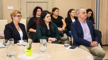 Grupa ludzi, mężczyzn i kobiet, siedzi w pomieszczeniu wokół stołu z filiżankami, szklankami i dzbankami na wodę. Są ubrani w strój biznesowy lub elegancki i wydają się uważni, prawdopodobnie słuchając mówcy na spotkaniu lub seminarium.