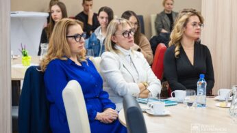 Grupa kobiet i mężczyzn siedzi przy stołach podczas imprezy w pomieszczeniu. Trzy kobiety na pierwszym planie noszą okulary i stroje biznesowe, patrząc uważnie przed siebie. Na stołach znajdują się kubki, szklanki, woda butelkowana i notatniki. Inni uczestnicy są widoczni w tle.