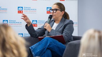 Kobieta w okularach, szarej marynarce i niebieskich dżinsach siedzi w szarym fotelu, trzyma mikrofon i teczkę, mówi i gestykuluje ręką. Za nią znajduje się biały baner z niebieskim i czerwonym logo Powiatu Krapkowickiego.