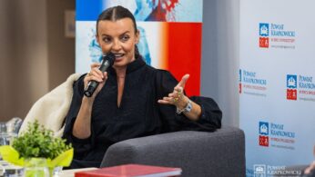 Kobieta z zaczesaną do tyłu fryzurą siedzi na szarym krześle, trzymając mikrofon i gestykulując lewą ręką. Ma na sobie czarny strój. W tle znajdują się banery z polskim tekstem i logo. Na pierwszym planie stół z dokumentami i rośliną.