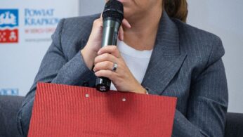 Kobieta w okularach i szarej marynarce siedzi trzymając mikrofon i czerwoną teczkę. Ma jasnobrązowe włosy zaczesane do tyłu i wygląda na zamyśloną. Za nią na szarej ścianie znajdują się banery Powiatu Krakowskiego. Scena sugeruje formalną dyskusję lub wydarzenie.