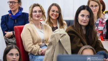 Grupa kobiet siedzi razem w jasno oświetlonym pokoju, niektóre uśmiechnięte i zaangażowane. Większość nosi luźne ubrania i okulary. Uwaga skupiona jest na kobiecie w centrum, która uśmiecha się szeroko, podczas gdy inne uważnie słuchają. W prawym dolnym rogu widoczne jest logo.