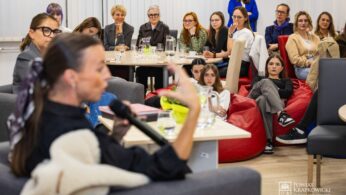 Grupa kobiet siedzi w dobrze oświetlonym pomieszczeniu, uważnie słuchając mówcy trzymającego mikrofon na pierwszym planie. Niektóre kobiety siedzą na krzesłach, inne na workach z fasolą. Na stołach stoją szklanki, dzbanki z wodą i notatniki.