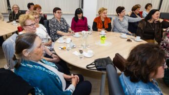 Grupa kobiet siedzi wokół okrągłych stołów z kubkami, talerzami i przekąskami, uważnie słuchając na spotkaniu w pomieszczeniu. Pokój ma wysokie żaluzje, a kobiety są ubrane w swobodne ubrania. W prawym dolnym rogu widoczne jest logo Powiatu Krakowskiego.