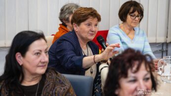 Kobieta w średnim wieku w niebieskiej kurtce trzyma mikrofon i gestykuluje podczas przemówienia w sali konferencyjnej. Jest otoczona przez inne kobiety, niektóre słuchają, a inne się uśmiechają. Otoczenie wydaje się formalne, ze szklanymi dzbankami i notatnikami na stole.