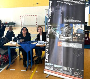 Dwie kobiety siedzą przy stoliku z broszurami w sali gimnastycznej, uśmiechając się do kamery. Obok nich znajduje się wysoki baner Zespołu Szkół Żeglugi Śródlądowej z obrazami działań morskich, informacjami o szkole i grafiką kotwicy. W tle znajdują się inne osoby.