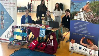 Grupa sześciu uczniów siedzi i stoi za kolorowym stołem wystawowym na szkolnych targach gimnastycznych. Na wystawie znajdują się banery, trofea, ulotki, czerwone bluzy i głowa manekina w okularach. Sprzęt sportowy i plakaty zdobią podłogę sali gimnastycznej i tło.