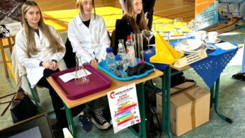 Cztery młode kobiety stoją na stoisku naukowym w sali gimnastycznej. Trzy siedzą za stołami z kolbami, zlewkami i papierami; dwie noszą białe fartuchy laboratoryjne. Jedna stoi za nimi, uśmiechając się. W tle widać banery, płaszcze, mężczyznę po prawej i bramkę do piłki ręcznej.