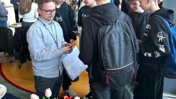 Mężczyzna w okularach i jasnej koszuli rozmawia z grupą nastoletnich chłopców ubranych w ciemne bluzy z kapturem i plecaki. Stoją obok stołu z różowymi tulipanami, broszurami i kubkiem kawy. W tle widoczne są inne osoby.