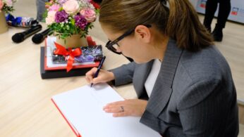 Kobieta z brązowymi włosami spiętymi w kucyk, w okularach i szarej marynarce, podpisuje książkę przy stole z kompozycjami kwiatowymi, stosem książek przewiązanych czerwoną wstążką i dwoma mikrofonami w pobliżu. Scena wygląda formalnie i profesjonalnie.