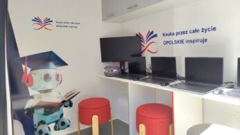 Nowoczesna klasa z komputerami i czerwonymi stołkami. Wyświetlacz ścienny przedstawia rysunkowego robota w czapce absolwenta trzymającego książkę. Na ścianach znajduje się polski tekst i kolorowe logo. Nad obszarem roboczym zamontowane są szafki, a światło słoneczne rozjaśnia pomieszczenie.