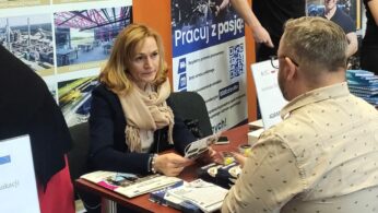 Kobieta i mężczyzna siedzą naprzeciwko siebie przy stole i rozmawiają. Kobieta trzyma broszury, ma na sobie płaszcz i szalik. Za nimi znajdują się plakaty z polskim tekstem, zdjęcia budynków i pracujących ludzi. Wygląda to na targi pracy lub wydarzenie związane z karierą.