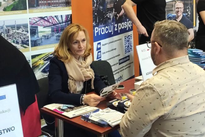 Kobieta i mężczyzna siedzą naprzeciwko siebie przy stole i rozmawiają. Kobieta trzyma broszury, ma na sobie płaszcz i szalik. Za nimi znajdują się plakaty z polskim tekstem, zdjęcia budynków i pracujących ludzi. Wygląda to na targi pracy lub wydarzenie związane z karierą.