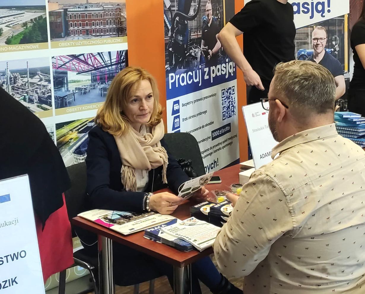 Kobieta i mężczyzna siedzą naprzeciwko siebie przy stole i rozmawiają. Kobieta trzyma broszury, ma na sobie płaszcz i szalik. Za nimi znajdują się plakaty z polskim tekstem, zdjęcia budynków i pracujących ludzi. Wygląda to na targi pracy lub wydarzenie związane z karierą.