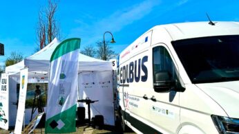 Biała furgonetka z napisem EDUBUS jest zaparkowana obok białego namiotu z banerami i składanymi krzesłami na zewnątrz. Niebo jest jasnoniebieskie, a podłoże wyłożone kostką brukową. W tle widoczne są wysokie, nagie drzewa i latarnia.