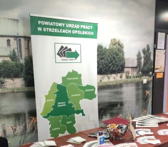 Stoisko z banerem przedstawiającym zieloną mapę regionów i napisem "POWIATOWY URZĄD PRACY W STRZELCACH OPOLSKICH". Na stole z przodu znajdują się foldery, broszury, miska z cukierkami i materiały biurowe. Z tyłu znajduje się malownicza fototapeta przedstawiająca nadrzeczną wioskę.