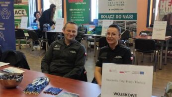 Dwóch umundurowanych funkcjonariuszy siedzi przy stoliku z broszurami, miską cukierków i medalami na targach pracy. Za nimi znajdują się banery pracodawców, w tym 