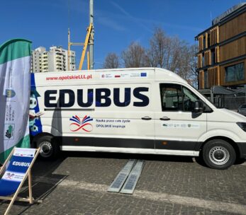 Biała furgonetka z napisem "EDUBUS" z edukacyjnymi banerami i logo zaparkowana na brukowanej ulicy. Nowoczesne budynki, drzewa bez liści i dźwigi budowlane są widoczne w tle pod czystym, błękitnym niebem.