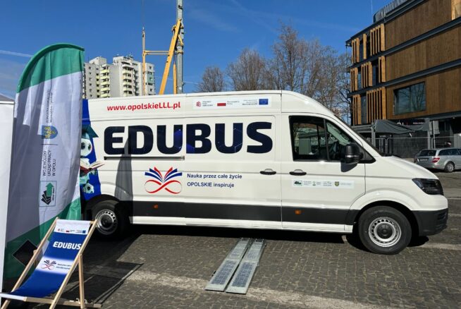 Biała furgonetka z napisem "EDUBUS" z edukacyjnymi banerami i logo zaparkowana na brukowanej ulicy. Nowoczesne budynki, drzewa bez liści i dźwigi budowlane są widoczne w tle pod czystym, błękitnym niebem.