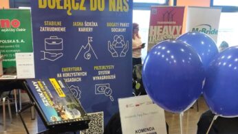 Stoisko rekrutacyjne lokalnej policji z niebieskim banerem z żółtymi napisami, broszurami na czarnym stojaku, niebieskimi balonami i napisem 