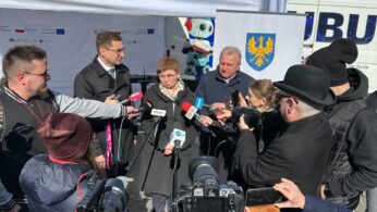 Kobieta przemawia do kilku mikrofonów podczas imprezy prasowej na świeżym powietrzu, otoczona przez dziennikarzy i kamerzystów. Stoi przed białym namiotem z banerami i niebiesko-żółtym herbem. Jest słonecznie, a ludzie noszą kurtki. W tle widać budynki i autobus.