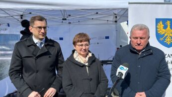 Trzy osoby stoją na zewnątrz przed białym namiotem z baldachimem i banerem z żółtym orłem. Dwóch mężczyzn ma na sobie ciemne płaszcze; kobieta w środku ma okulary i szary płaszcz. Jeden z mężczyzn mówi do mikrofonu na statywie. Niebo jest czyste i słoneczne.