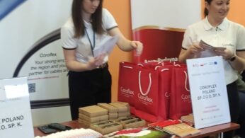 Dwie kobiety stoją za stołem na imprezie wewnętrznej, rozdając przedmioty. Na stole znajdują się czerwone torby Coroflex, broszury, małe pudełka i długopisy. Za nimi wyświetlane są banery Coroflex i Agro Rydzyna. Z przodu stoi kilka pustych krzeseł.