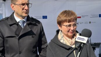 Mężczyzna w ciemnym trenczu i okularach stoi obok kobiety w czarnym płaszczu i szaliku. Oboje znajdują się na zewnątrz przed białym namiotem z logo i tekstem. Kobieta mówi do mikrofonu z napisem telewizja tv. Jest słonecznie.