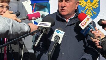 Mężczyzna w marynarce przemawia do kilku mikrofonów trzymanych przez reporterów. Stoi na zewnątrz w pobliżu baneru przedstawiającego żółtego orła na niebiesko-białym tle, prawdopodobnie godło regionu. Inni ludzie i zadaszenie namiotu są widoczne w tle w jasnym świetle słonecznym.