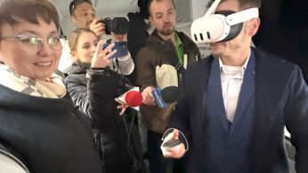 Mężczyzna w garniturze nosi biały zestaw słuchawkowy wirtualnej rzeczywistości i trzyma kontroler, podczas gdy wokół niego tłoczy się kilka osób z kamerami i mikrofonami. Po lewej stronie uśmiecha się kobieta w okularach i szaliku. Wygląda na to, że grupa znajduje się wewnątrz pojazdu lub małego pomieszczenia.