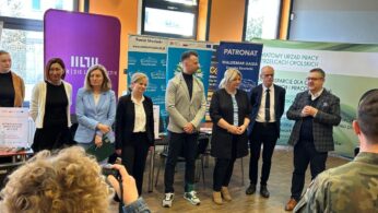 Grupa mężczyzn i kobiet stoi w pomieszczeniu przed banerami na imprezie. Jeden mężczyzna w kraciastej marynarce przemawia, gestykulując ręką. Inni uważnie słuchają. Na pierwszym planie siedzą ludzie. Pomieszczenie ma pomarańczowe i zielone ściany oraz duże okna.