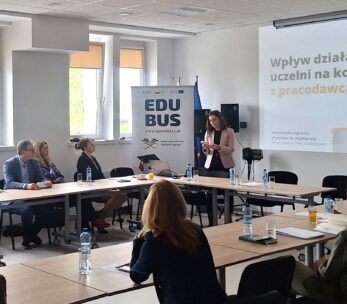 Kobieta stoi i przemawia do grupy osób siedzących wokół stołu konferencyjnego w jasnym pomieszczeniu. Na slajdzie prezentacji widnieje napis Wpływ działania uczelni na kontakt z pracodawcami. Na stole leży woda butelkowana i dokumenty, a na banerze widnieje napis EDUBUS.