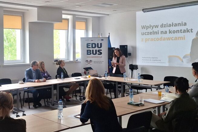 Kobieta stoi i przemawia do grupy osób siedzących wokół stołu konferencyjnego w jasnym pomieszczeniu. Na slajdzie prezentacji widnieje napis Wpływ działania uczelni na kontakt z pracodawcami. Na stole leży woda butelkowana i dokumenty, a na banerze widnieje napis EDUBUS.