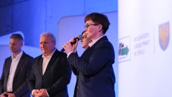 Cztery osoby w strojach formalnych stoją na scenie. Osoba z przodu, trzymająca mikrofon, mówi, podczas gdy pozostali słuchają. Tło jest podświetlone na niebiesko z białą ścianą z logo i emblematami, co sugeruje formalne wydarzenie lub prezentację.