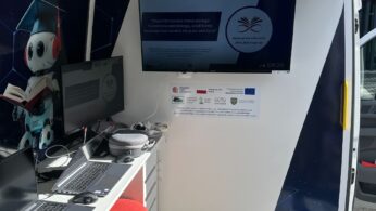 Wewnątrz mobilnej klasy o nazwie EDUBUS znajdują się laptopy na biurku, trzy okrągłe stołki, telewizor wyświetlający treści edukacyjne oraz plakat ścienny przedstawiający robota w czapce absolwenta ze słuchawkami. Światło słoneczne wpada przez otwarte drzwi.