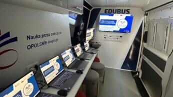Obraz przedstawia wnętrze mobilnej klasy o nazwie EDUBUS, z rzędem otwartych laptopów na długim biurku, z których każdy wyświetla ten sam slajd edukacyjny. Przed laptopami znajdują się czerwone, wyściełane stołki, a z przodu duży ekran prezentacyjny.
