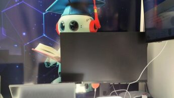 Kreskówkowy robot w niebieskiej czapce absolwenta i czerwonych słuchawkach trzyma otwartą książkę na cyfrowym tle. Z przodu znajduje się laptop, duży monitor częściowo zasłaniający robota, słuchawki VR i kable na jasnym biurku.