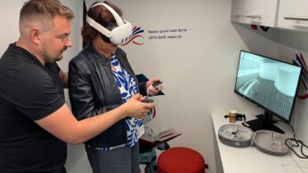 Mężczyzna pomaga kobiecie noszącej gogle VR, podczas gdy ona trzyma kontrolery. Stoją w małym, białym pokoju z czerwonym stołkiem i monitorem komputerowym wyświetlającym wirtualne środowisko. W pobliżu na stole leży sprzęt VR. Na ścianie widoczny jest polski tekst.