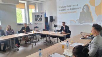 Grupa osób w strojach biznesowych siedzi wokół stołu konferencyjnego w kształcie litery U i dyskutuje. Slajd prezentacji na ścianie głosi Wpływ działania uczelni na kontakt z pracodawcami. Przy oknach stoją flagi i transparenty. Na stole leżą butelki wody i gazety.