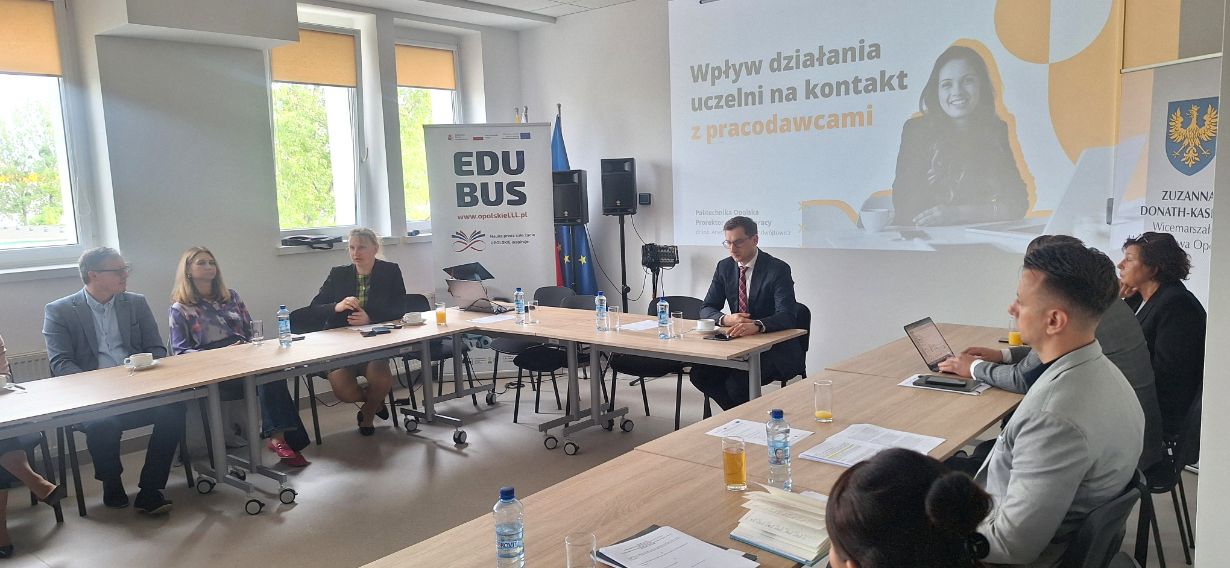 Grupa osób w strojach biznesowych siedzi wokół stołu konferencyjnego w kształcie litery U i dyskutuje. Slajd prezentacji na ścianie głosi Wpływ działania uczelni na kontakt z pracodawcami. Przy oknach stoją flagi i transparenty. Na stole leżą butelki wody i gazety.