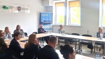 Sala konferencyjna z 12 osobami, zarówno mężczyznami, jak i kobietami, siedzącymi wokół trzech stołów w kształcie litery 