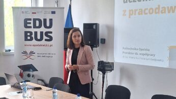 Kobieta w jasnoróżowej marynarce stoi w pobliżu stołu konferencyjnego z butelkami wody, krzesłami i papierami. Za nią znajduje się ekran projektora z polskim tekstem i stojący baner z napisem EDUBUS. Pokój ma duże okna i jasne oświetlenie.