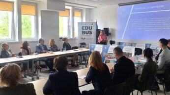 Kobieta stoi i prezentuje slajdy grupie osób siedzących przy stołach konferencyjnych w jasnym pomieszczeniu. Slajd na ekranie brzmi 